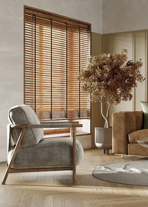Veneciana de madera color miel con cintas oscuras en un salón con butaca gris de brazos de madera y un árbol en maceta