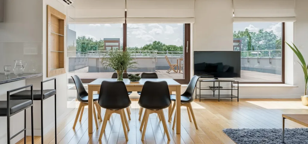Salón comedor con estores paqueto claros en grandes ventanales, mesa de madera y sillas negras