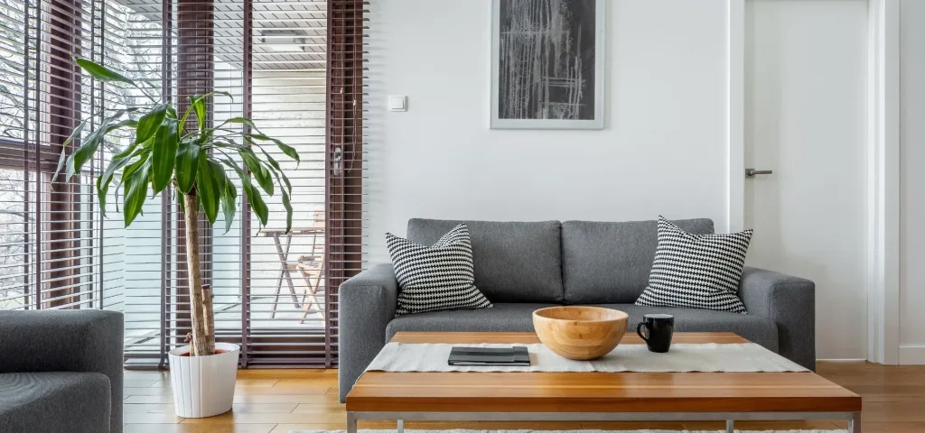 Venecianas de madera oscura en los ventanales de un salón con sofá gris y mesa de centro de madera