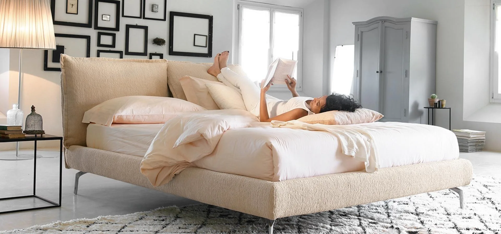 Cama tapizada en beige con patas metálicas y cabecero bajo, con una mujer leyendo sobre ella
