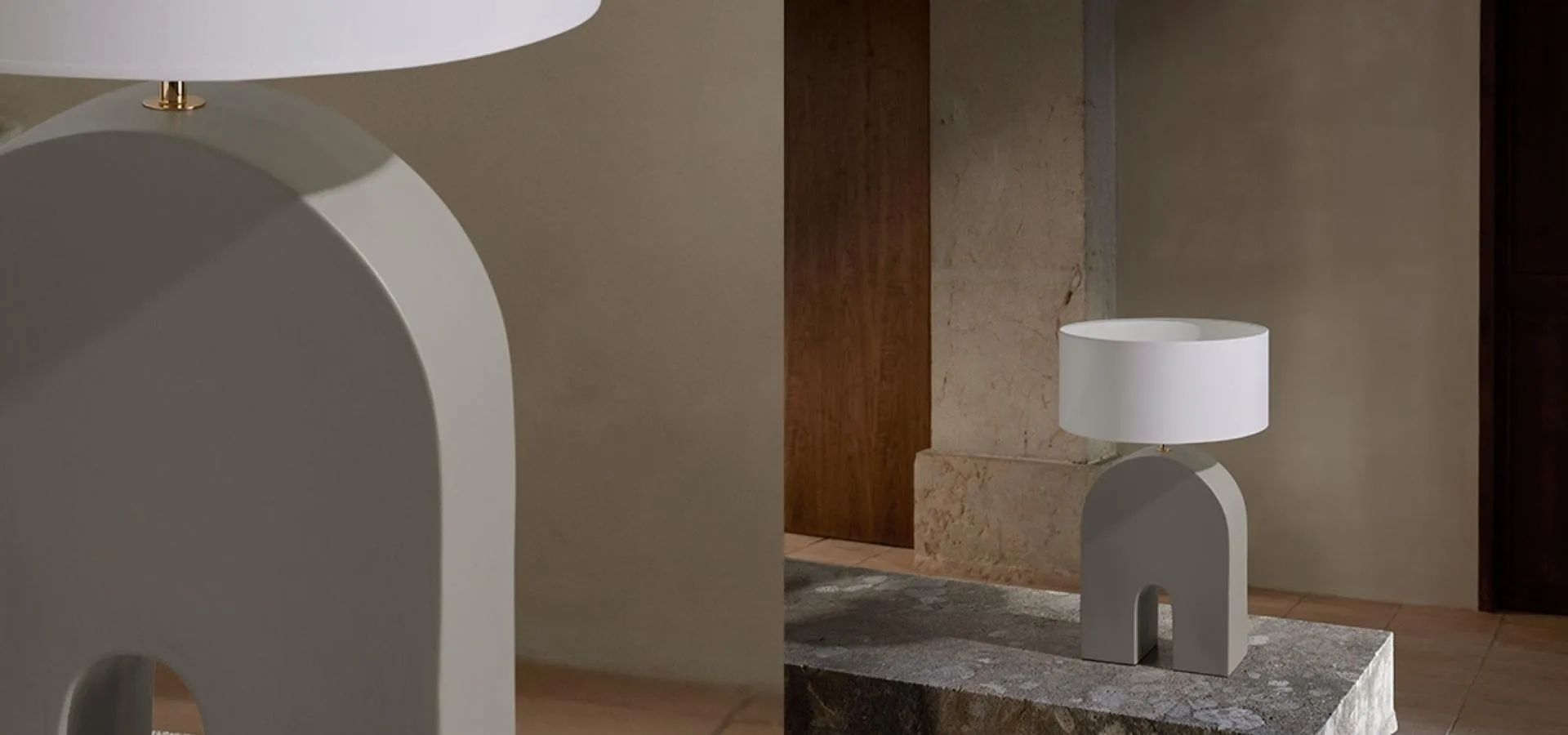 Lámpara de mesa con pie gris en forma de arco y pantalla blanca, en detalle y sobre una mesa veteada