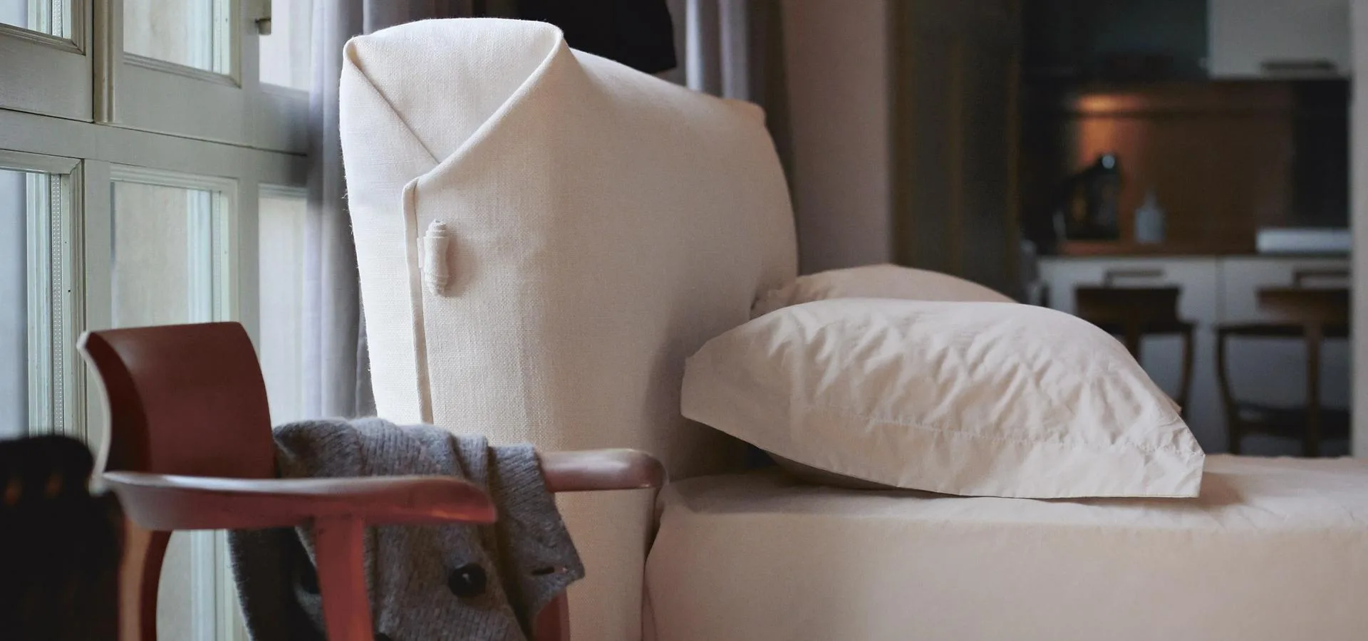 Cabecero tapizado en blanco con funda de solapa y botón, junto a una silla de madera y una almohada