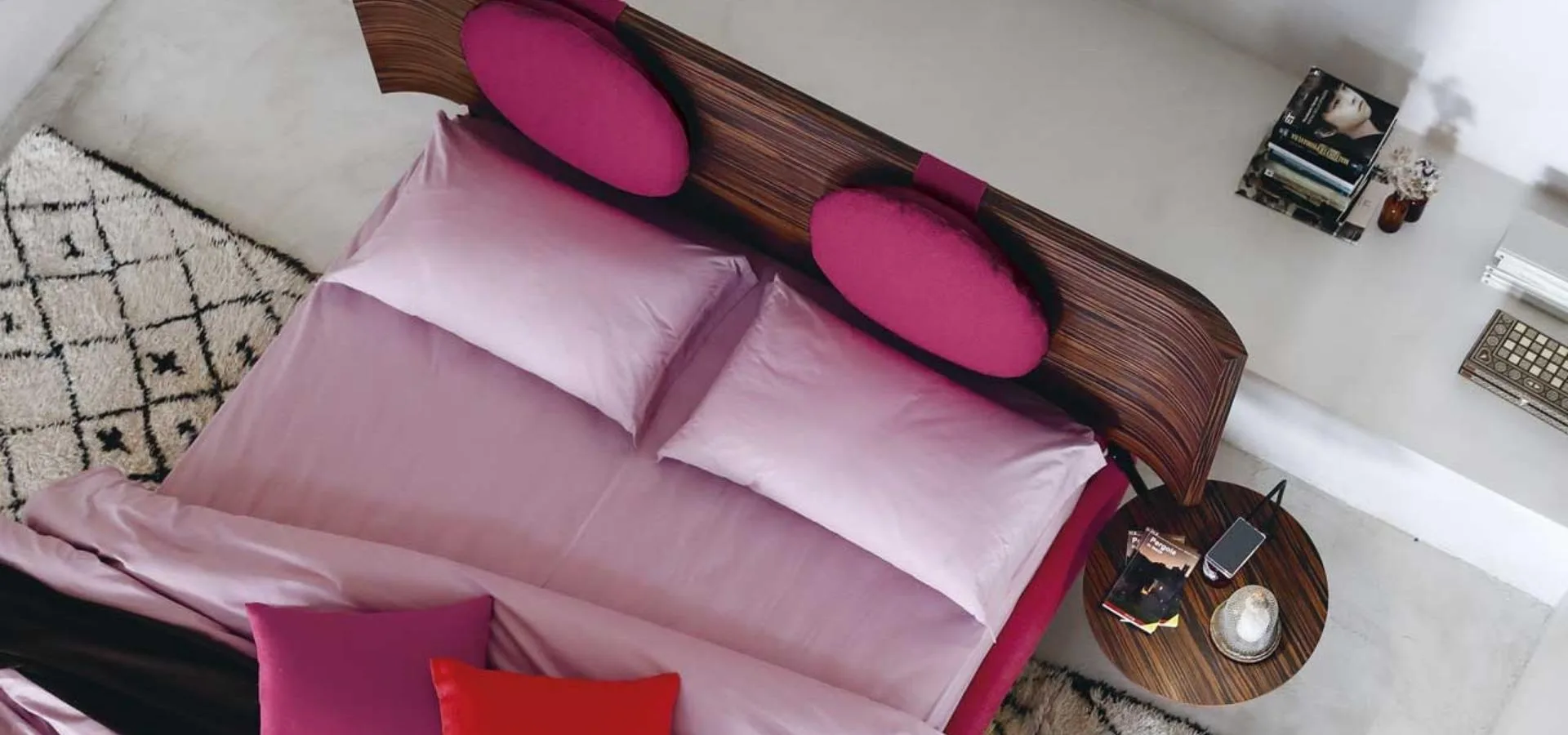 Cama vista desde arriba con ropa de cama rosa y fucsia y cabecero de madera con dos cojines redondos