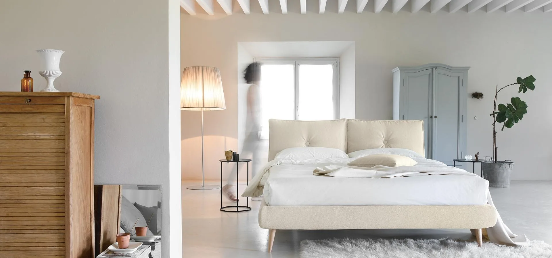 Cama de cabecero acolchado en crema en un dormitorio blanco con cómoda de madera, lámpara y planta