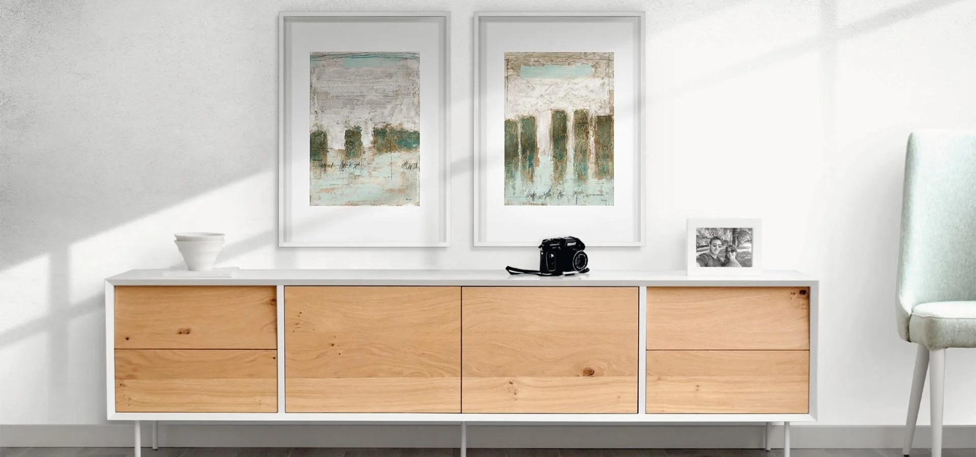 Dos cuadros abstractos en verde y gris con marco blanco sobre un aparador blanco y madera clara