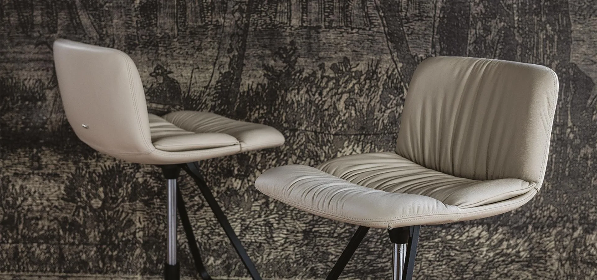 Dos sillas tapizadas en beige con respaldo de pliegues y base metálica oscura frente a una pared de textura gris