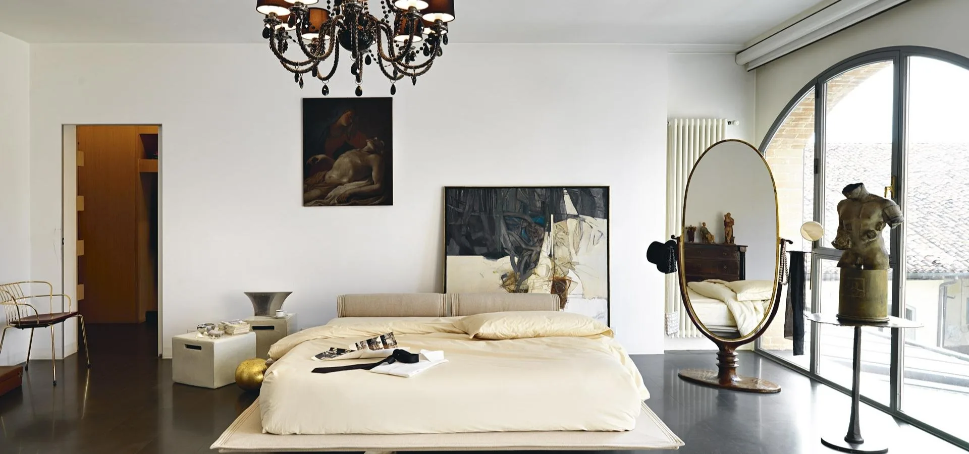 Cama baja con ropa de cama blanca en un dormitorio amplio con lámpara de araña y espejo de pie ovalado