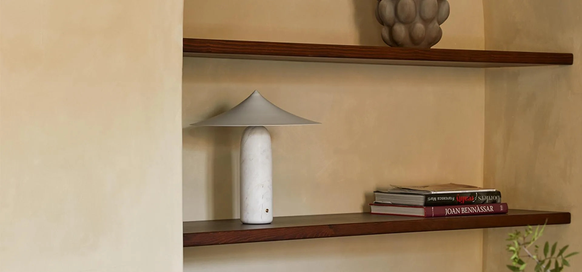 Lámpara de mesa blanca con pantalla cónica sobre una balda de madera oscura, junto a libros apilados