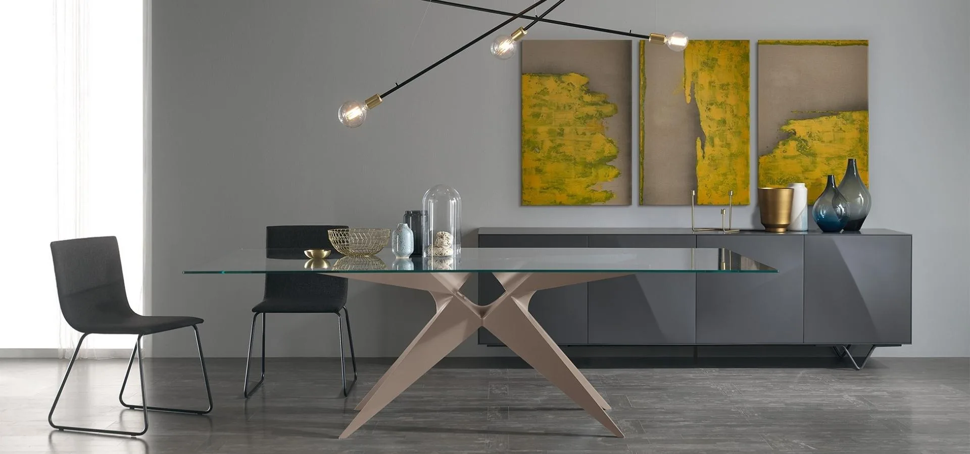 Comedor con mesa de cristal y patas de madera cruzadas, sillas negras y tres lienzos abstractos amarillos