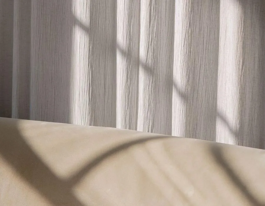 Luz del sol a través de un visillo de pliegues verticales, con sombras sobre una superficie clara