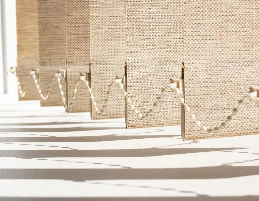 Detalle de lamas verticales en tejido beige de trama gruesa, con la cadena inferior y sombras al sol