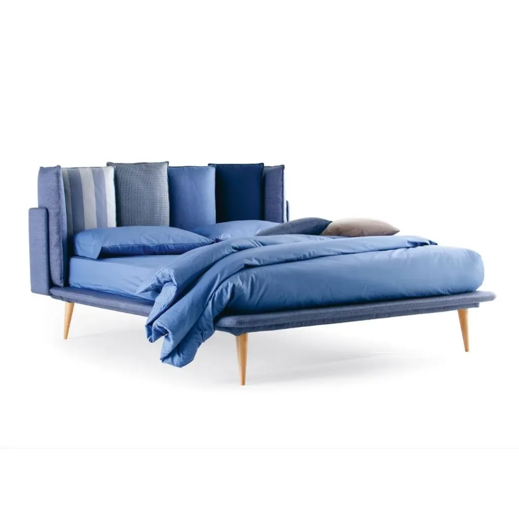 Cama tapizada en azul con cabecero de cojines en varios tonos de azul y patas de madera inclinadas