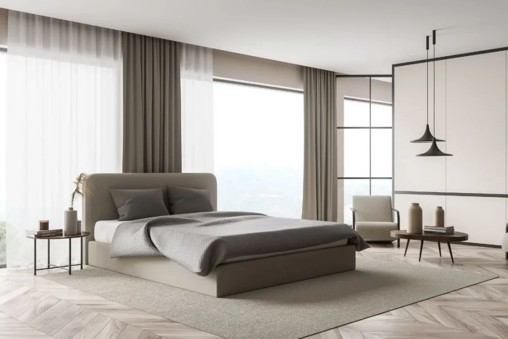 Dormitorio con cama tapizada gris, cortinas grises y visillos blancos en los ventanales