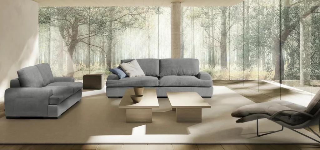 Salón con dos sofás grises, mesas bajas de madera clara y chaise longue frente a un ventanal al bosque